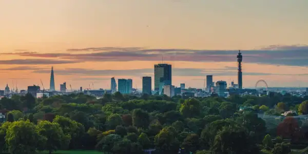 London im Sommer: Die Skyline mit dem Shard und dem BT Tower ragt über einem grünen Park auf, während die Sonne den Himmel in warme Farben taucht.