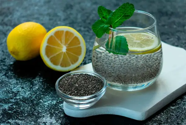 Ett glas med chia vatten, garnerat med citron och mynta. Bredvid står en skål med chiafrön och två citroner.