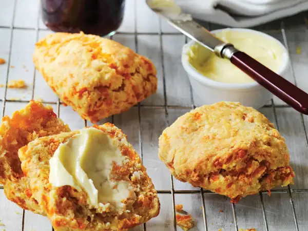 Trois scones américains dorés, dont un coupé en deux et garni de beurre, sur une grille. Un pot de confiture et du beurre à tartiner sont à proximité.