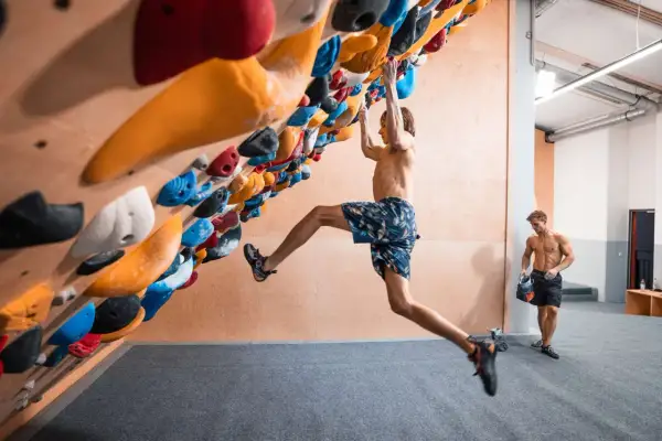 Ein Mann klettert an einer Boulderwand, während ein anderer Mann daneben steht. Der Unterschied zwischen Klettern und Bouldern wird hier deutlich: keine Seile, nur Griffe und eine Matte.