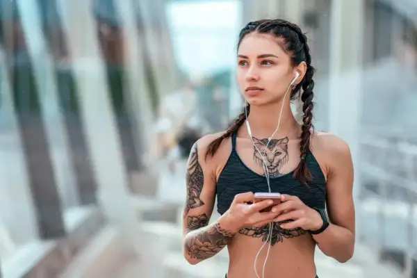 Kobieta z warkoczami i słuchawkami na uszach, trzymająca telefon. Widoczny delikatny tatuaż między piersiami w kształcie kota.