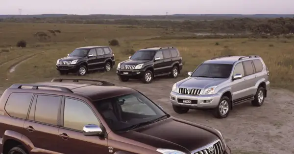 Cztery samochody Toyota Land Cruiser 120, w tym jeden brązowy, dwa czarne i jeden srebrny, stoją na polnej drodze wśród traw.