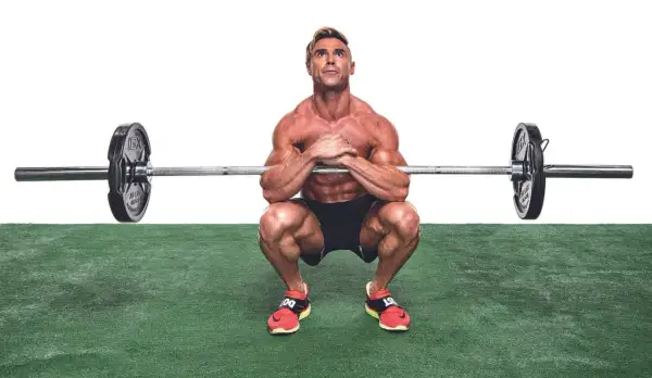 Muskulös man utför en zercher squat med skivstång i armvecken.