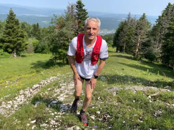 Un coureur en tenue de trail, sac à dos rouge, sur un sentier escarpé près d'Oyonnax, profitant d'un paysage verdoyant et vallonné.