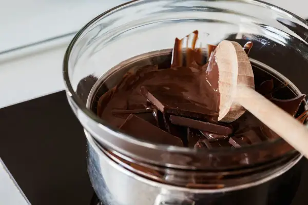 Trozo de chocolate oscuro derritiéndose en baño maría, con una cuchara de madera. Así es como derretir chocolate para postres.