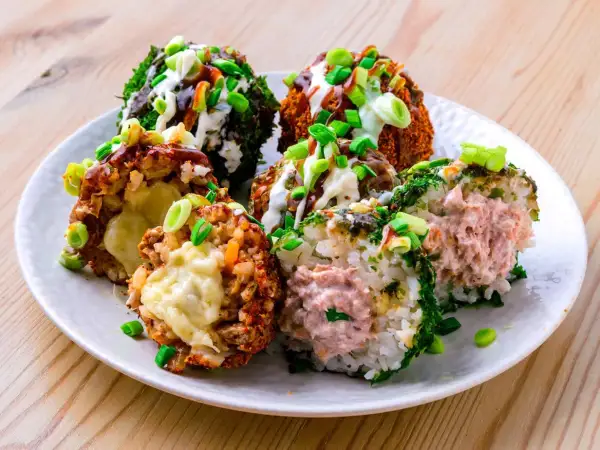 Sechs bunte Onigiri mit verschiedenen Füllungen und Toppings, umgeben von Frühlingszwiebeln und Saucen.