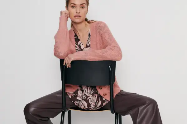 Elegancka kobieta w różowym swetrze Gerry Weber i brązowych spodniach z ekoskóry siedzi na czarnym krześle.