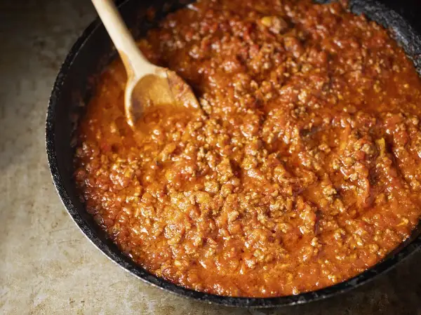 Un saporito ragù di carne in una padella, pronto per essere servito. Un cucchiaio di legno mescola la salsa rossa e densa.
