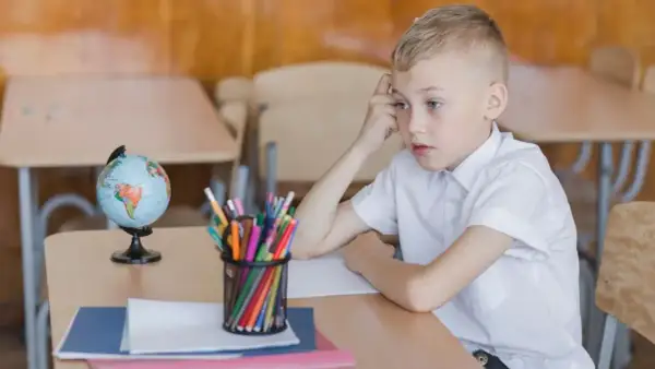 Chłopiec w białej koszuli siedzi przy biurku z globusem i kredkami. Zamyślony, może czeka na odroczenie obowiązku szkolnego.