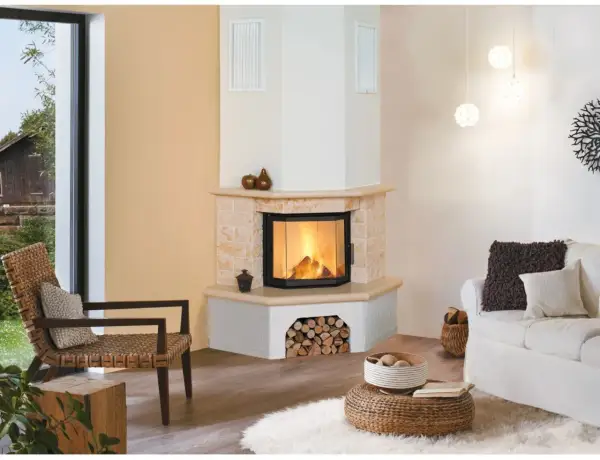 Gemütliches Wohnzimmer mit einem Eck-Kachelofen, der einladend brennt. Ein Rattansessel und ein weißes Sofa laden zum Entspannen ein.