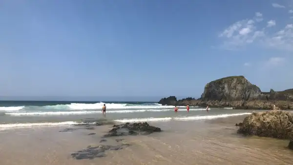 Gente disfrutando del mar en la playa de Frouxeira, con olas rompiendo y una gran roca verde al fondo bajo un cielo azul.