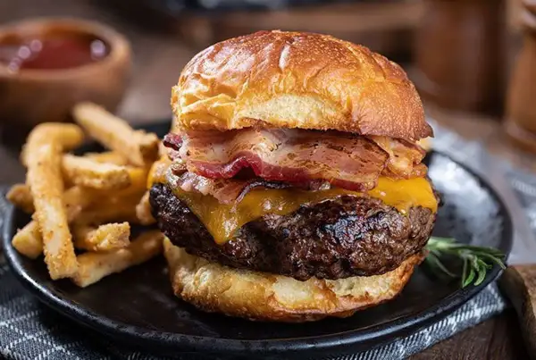 Gros plan sur un burger maison avec bacon, fromage fondant et frites croustillantes sur une assiette noire.