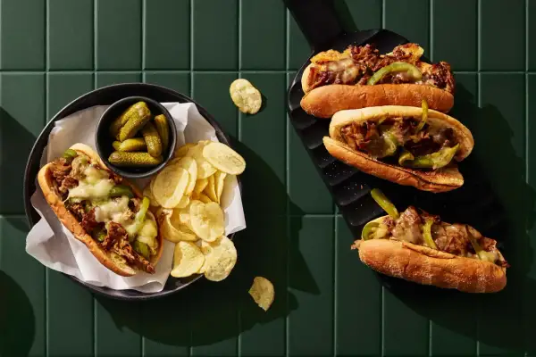 Två philly cheese steak-mackor med smält ost, paprika och lök. Serveras med cornichoner och potatischips.