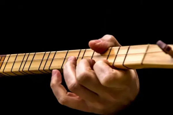 Dłoń gra na instrumencie, dociskając struny. Widoczne są progi i nazwy strun w gitarze.