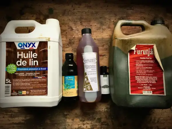 Plusieurs bouteilles d'huile pour le bois, dont de l'huile de lin et du goudron de pin, sont alignées sur une surface en bois.