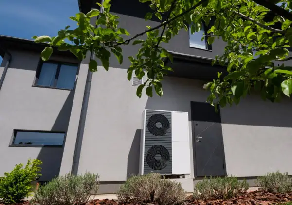 Unità esterna di una pompa di calore residenziale, moderna e discreta, installata accanto a una porta d'ingresso su un edificio grigio.