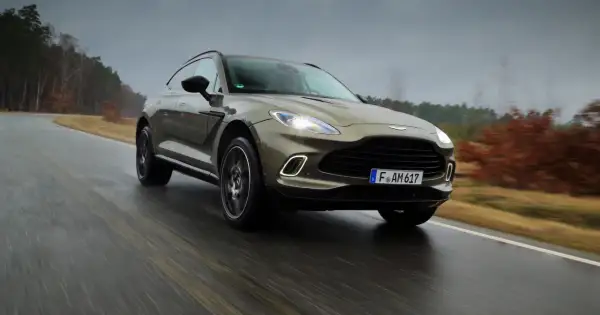 Elegancki Aston Martin SUV przemierza mokrą drogę wśród jesiennych drzew.