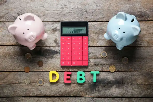 Due salvadanai rosa e azzurro, una calcolatrice rossa con un numero elevato e lettere che formano la parola "DEBT" suggeriscono un **debito residuo** da gestire.