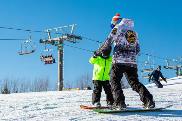Instruktor snowboardingu uczy dziecko na stoku, w tle kolejka krzesełkowa.