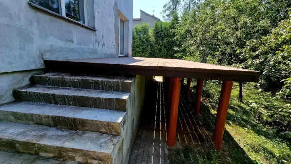 Nowy taras na słupach stalowych, z drewnianą podłogą, sąsiaduje z betonowymi schodami i zielonym ogrodem.