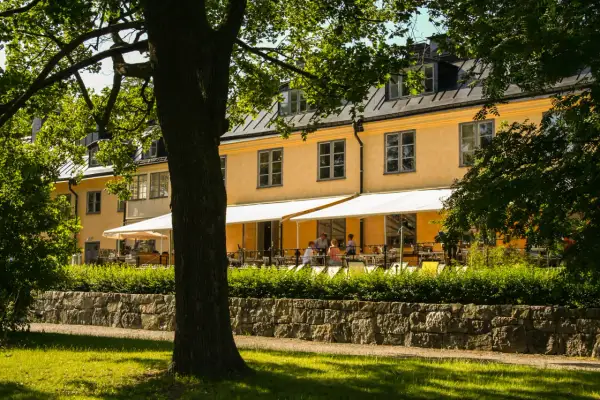 En gul byggnad med vita markiser och uteservering. En lång rad med träd skymmer delvis vyn.