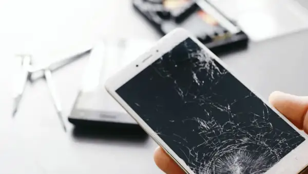 Dłoń trzyma telefon z pękniętym ekranem. Obok leżą narzędzia do naprawy.