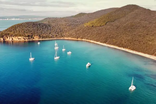 Vela al largo della costa della Maremma Toscana. Il mare azzurro e cristallino abbraccia una baia con diverse barche a vela e un catamarano.