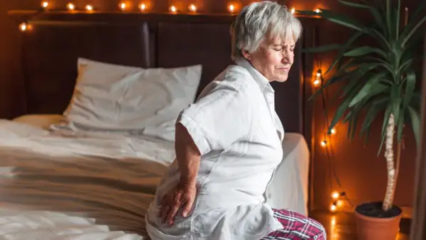Elderly woman grimaces in pain, holding her hip. She looks like she slept on her hip wrong, sitting on a bed with festive lights.