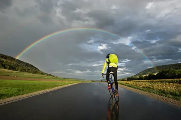 Rennradtour 3 Tage: Ein Radfahrer fährt unter einem Regenbogen auf nasser Straße.