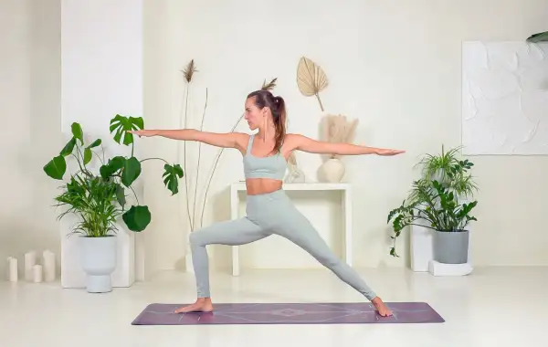 Mujer en postura de Virabhadrasana 2, con brazos extendidos y mirada al frente, rodeada de plantas y decoración minimalista.