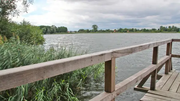 Drewniany pomost nad jeziorem, gdzie można łowić ryby. W oddali widać zalesiony brzeg i domy.