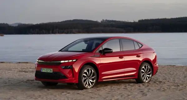 Czerwony Skoda Enyaq na piaszczystym brzegu, z lodowym jeziorem i lasem w tle.