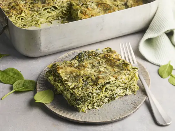 Eine Portion Spinat Ricotta Lasagne auf einem Teller, daneben eine Auflaufform mit der restlichen Lasagne. Frische Spinatblätter liegen daneben.