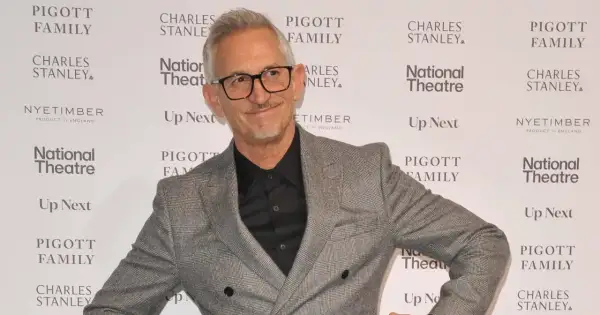 Gary Lineker w szarej marynarce i okularach, uśmiecha się na tle ścianki z logo National Theatre.