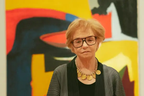 A woman with glasses and a statement necklace stands before a vibrant abstract expressionist painting.