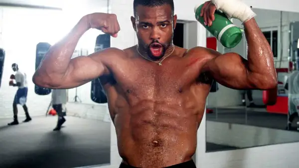 A sweaty boxer flexes his biceps in a gym, holding a green water bottle. He's training hard, ready for any division, perhaps even the Roy Jones Jr. division.