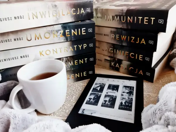 Stos książek Remigiusza Mroza, biały kubek z herbatą i czytnik Kindle.