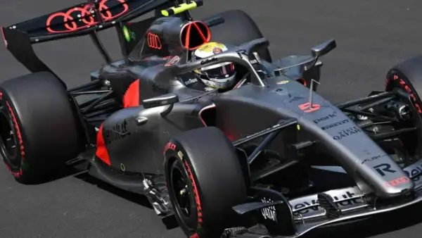 Un coche de **Audi F1** gris y negro con detalles rojos acelera en la pista. El piloto lleva casco amarillo y blanco.