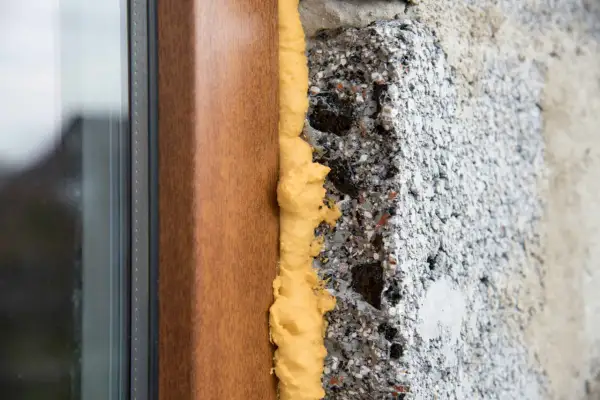 Dettaglio di un telaio di finestra in legno con schiuma poliuretanica espansa che sigilla lo spazio tra il telaio e il muro grezzo. Questo mostra come modellare la schiuma poliuretanica per un isolamento efficace.