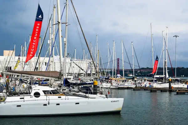 Una **barca lunga e sottile**, la Scarlett, è ormeggiata in un porto pieno di altre barche a vela. Sullo sfondo, un ponte rosso attraversa il fiume.