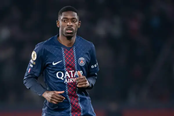 Piłkarz PSG, gdzie gra Dembele, w ciemnoniebieskiej koszulce z czerwonymi i białymi detalami, na tle rozmytego stadionu.