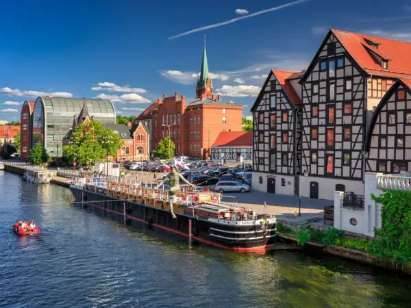 Malownicze stare miasta w Polsce nad rzeką. Barka z rzeźbą, łódka z ludźmi, zabytkowe kamienice i nowoczesna architektura.