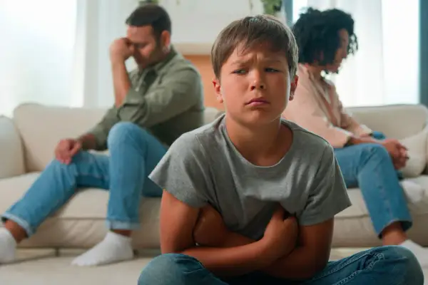 Bambino triste in primo piano, con i genitori separati seduti alle sue spalle, voltati l'uno dall'altra.