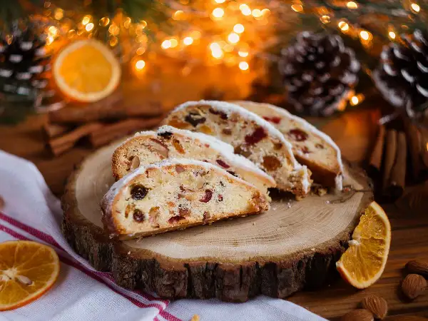 Tranches de stollen cake, un délice de Noël aux fruits secs et noix, saupoudré de sucre glace, sur une planche de bois, entouré de décorations festives.