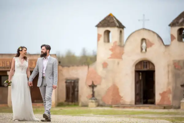 Un couple rayonnant, tout juste marié, se promène main dans la main devant une chapelle rustique, évoquant un mariage western.