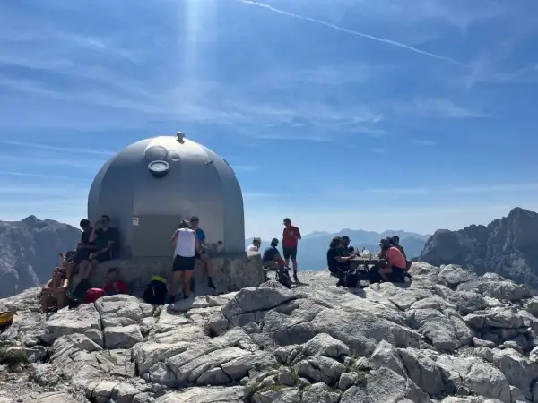 Gente descansa junto a un refugio en un viaje a Picos de Europa. El sol brilla en un cielo azul con nubes.