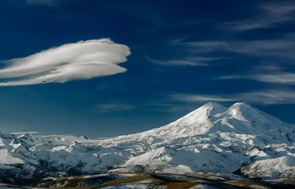 Pokryty śniegiem Elbrus, najwyższy szczyt Rosji, góruje nad ośnieżonymi pasmami górskimi pod błękitnym niebem z charakterystyczną chmurą.