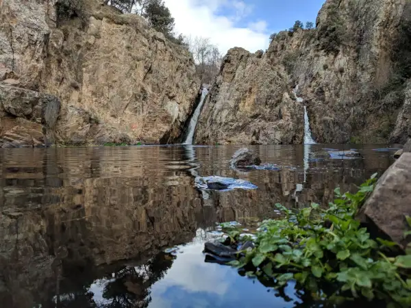 Cascadas gemelas caen entre rocas escarpadas en el embalse de Pedrezuela, reflejándose en el agua tranquila.