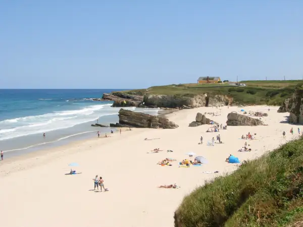 Gente disfrutando del sol y el mar en la playa Longara Barreiros, con formaciones rocosas y una casa amarilla al fondo.