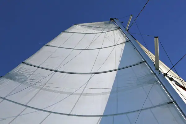 Vela bianca randa full batten spiegata al vento, con stecche orizzontali e cavi che salgono verso il cielo azzurro.